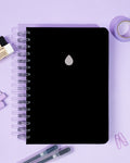 A black A5 notebook with a silver foiled raindrop logo on a purple background with stationery items.