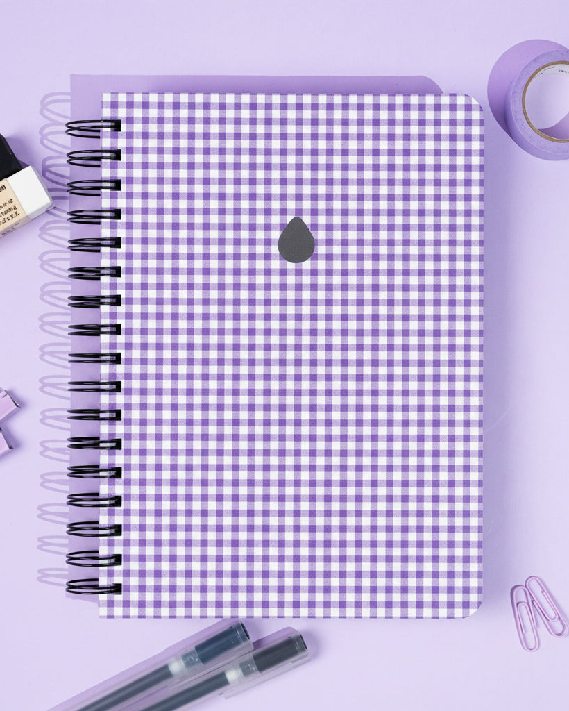 An A5 notebook with purple gingham design on a purple background with stationery items.