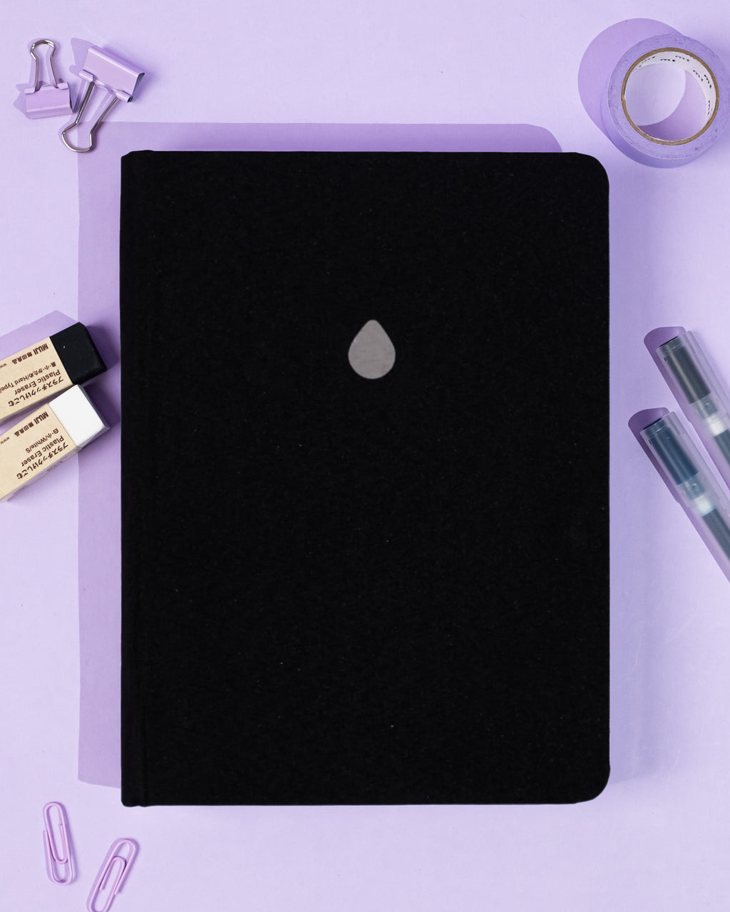 Black notebook with a silver logo on a purple background with stationery items.