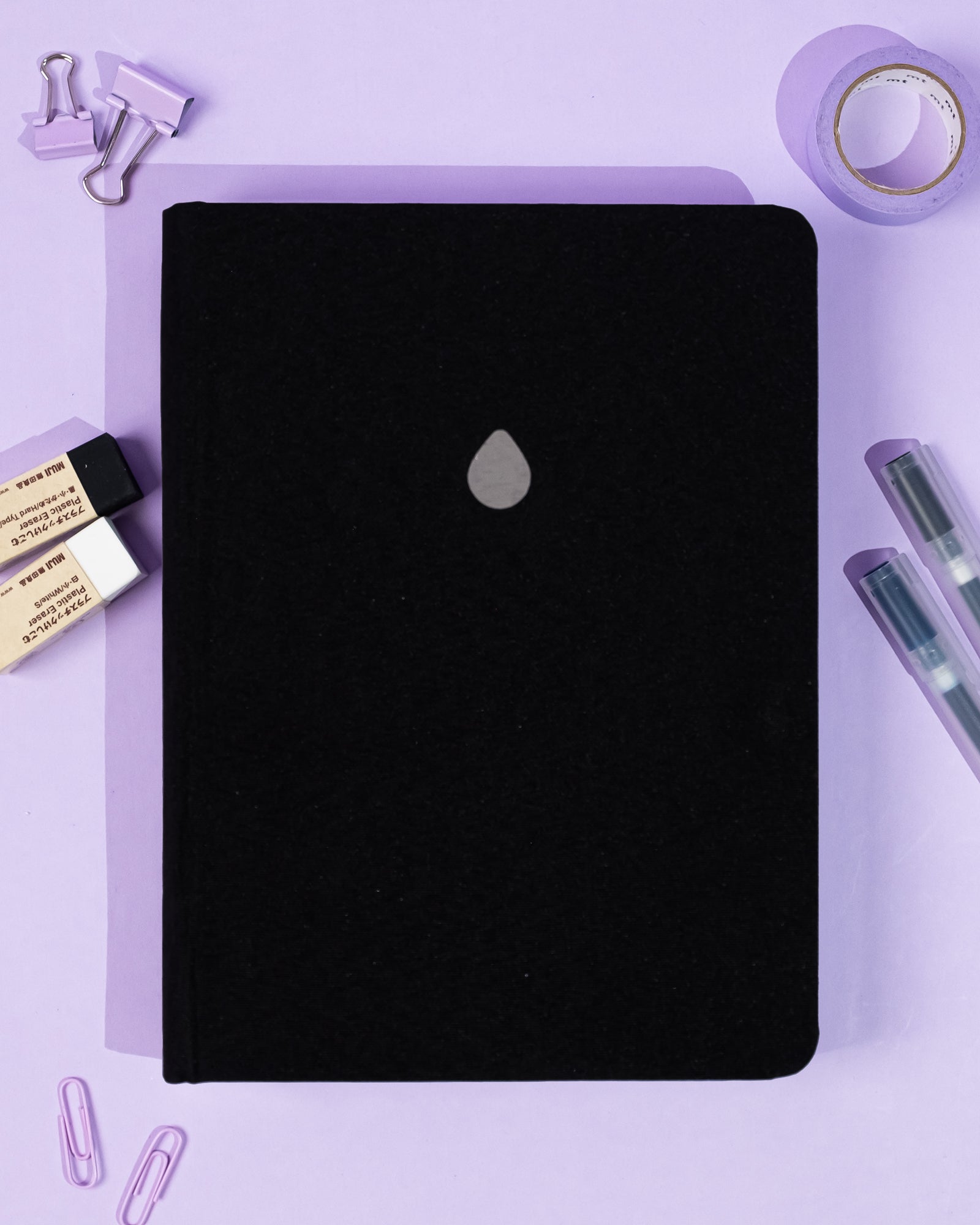 Black notebook with a silver logo on a purple background with stationery items.