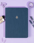 Denim blue notebook with a silver drop logo on a purple background with stationery items.