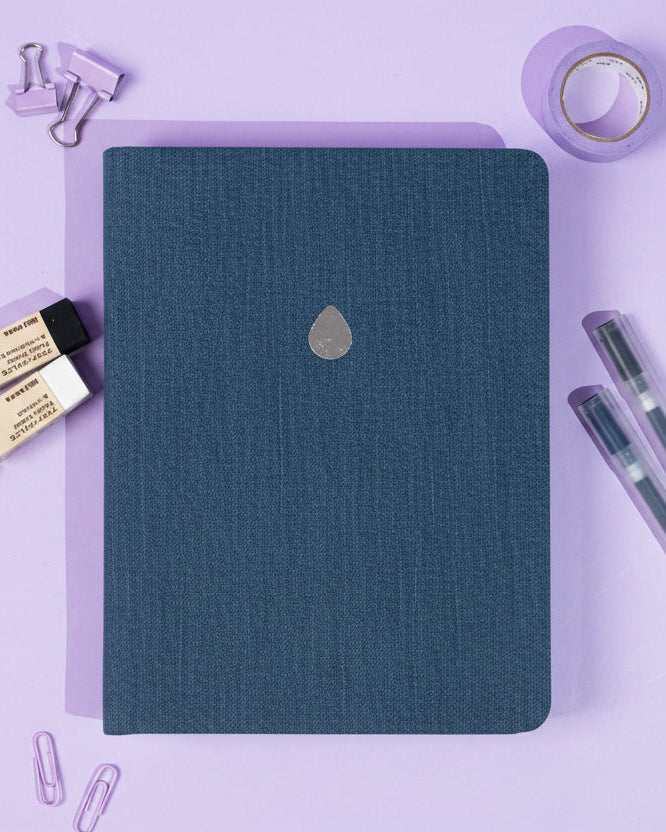 Denim blue notebook with a silver drop logo on a purple background with stationery items.